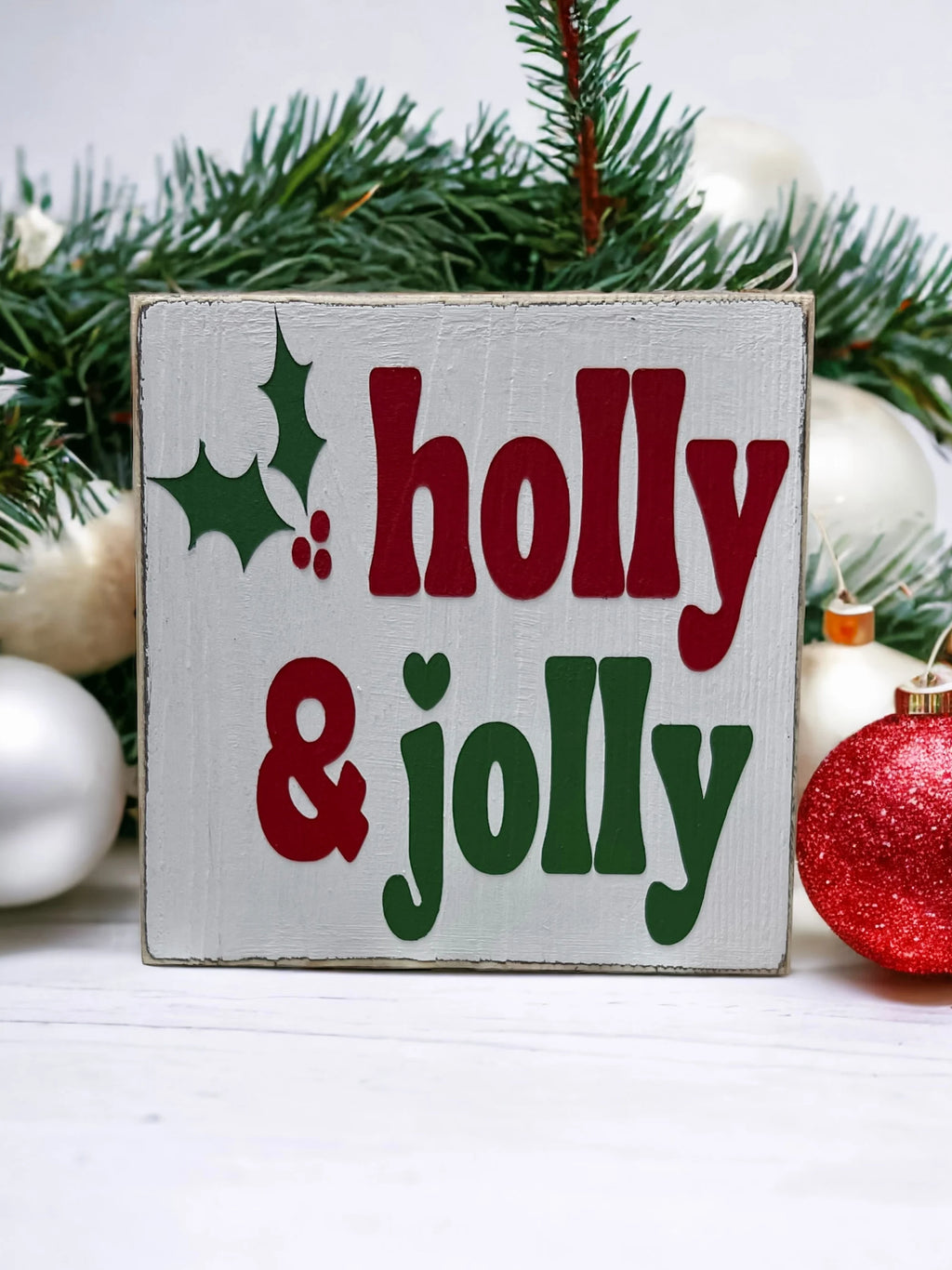Holly & Jolly wood sign with red ornaments and pine cone on white wood surface
