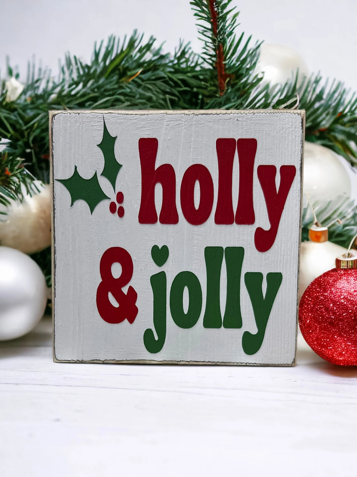 Holly & Jolly wood sign with red ornaments and pine cone on white wood surface