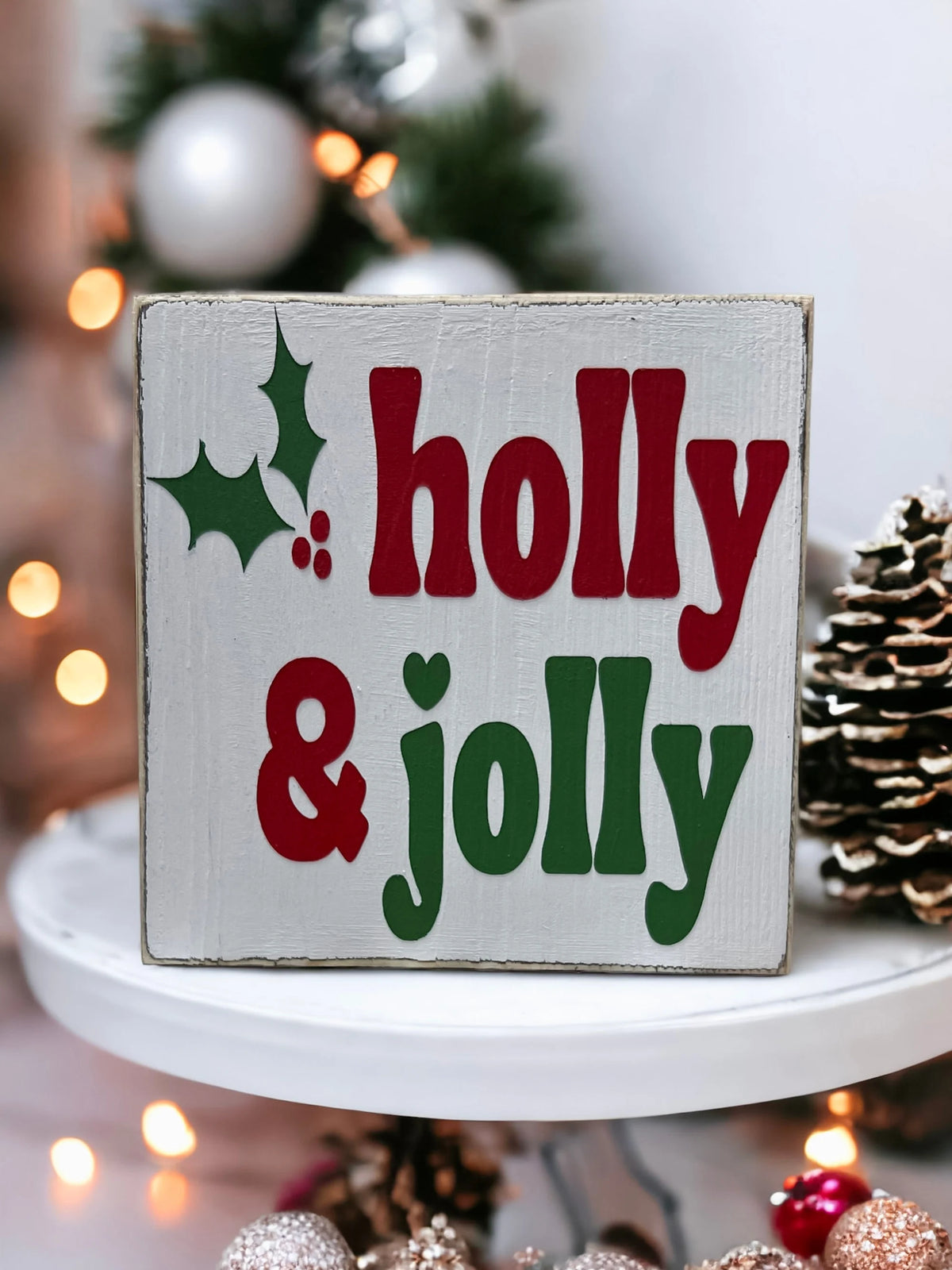 Holly & Jolly wood sign with pine cones and red ornament on white surface