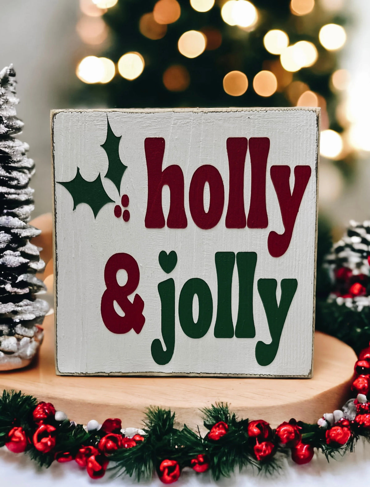 Holly & Jolly wood sign with pine garland and mixed ornaments on white surface