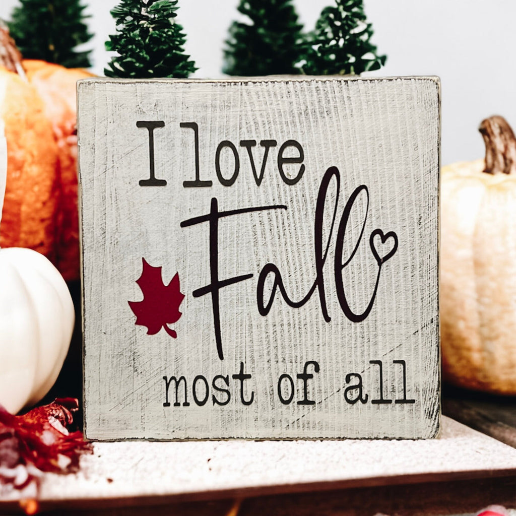 I Love Fall Most of All distressed off-white wood block sign with black lettering and burgundy maple leaf on white cake stand with orange pumpkins and pine cones hero shot