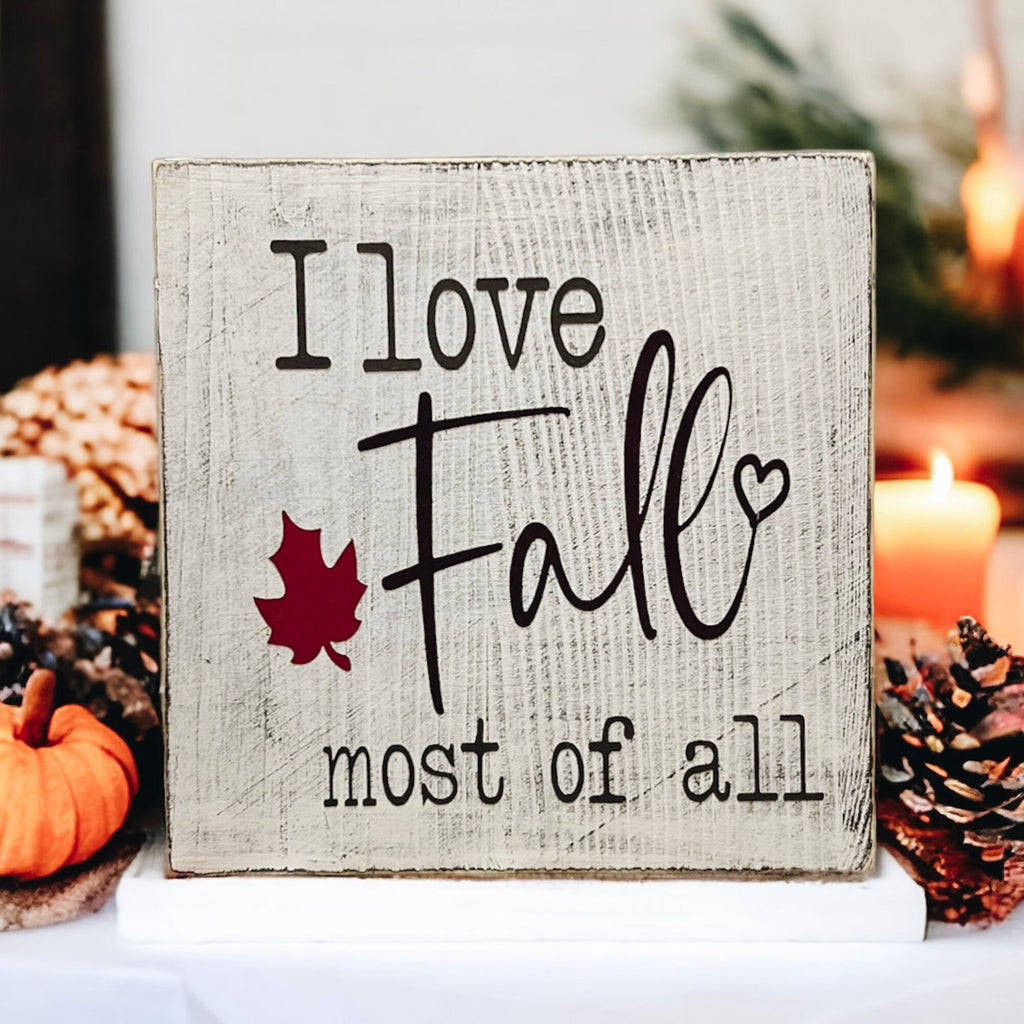 I Love Fall Most of All distressed off-white wood block sign with black lettering and burgundy maple leaf on white plate with orange pine cones and dried botanicals close up