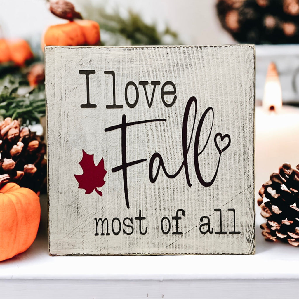 I Love Fall Most of All distressed off-white wood block sign with black lettering and burgundy maple leaf on white cake stand with orange pumpkins and pine cones side angle