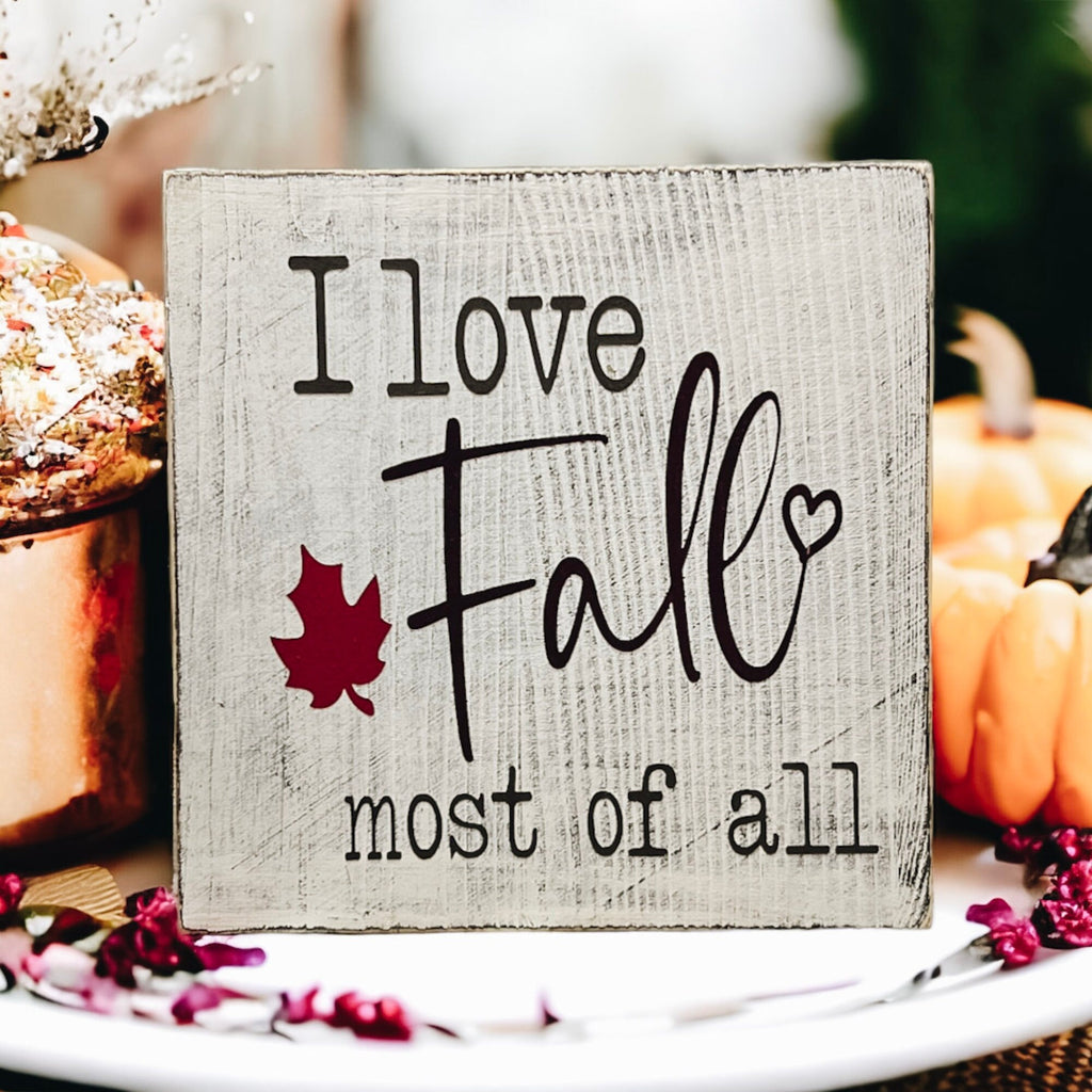 I Love Fall Most of All distressed off-white wood block sign with black lettering and burgundy maple leaf on white cake stand with orange pumpkins and pine cones angled view