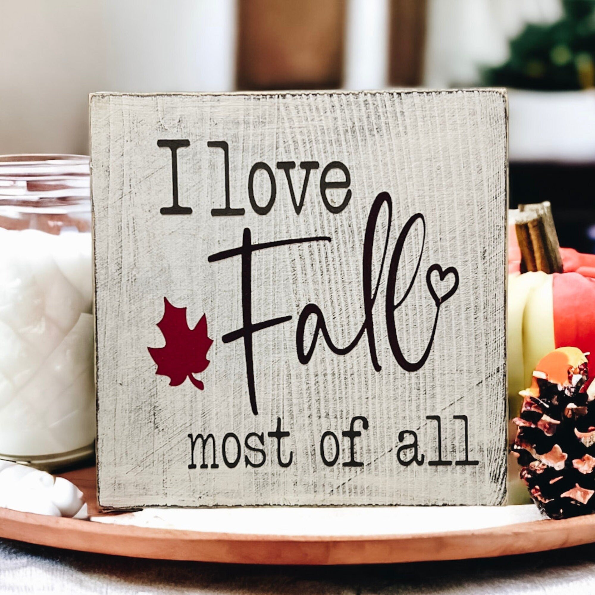 I Love Fall Most of All distressed off-white wood block sign with black lettering and burgundy maple leaf on white plate with orange pine cones and dried botanicals