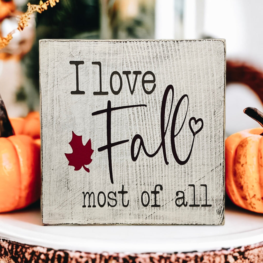 I Love Fall Most of All distressed off-white wood block sign with black lettering and burgundy maple leaf on white surface with pine cones orange pumpkins and lit candles wide shot