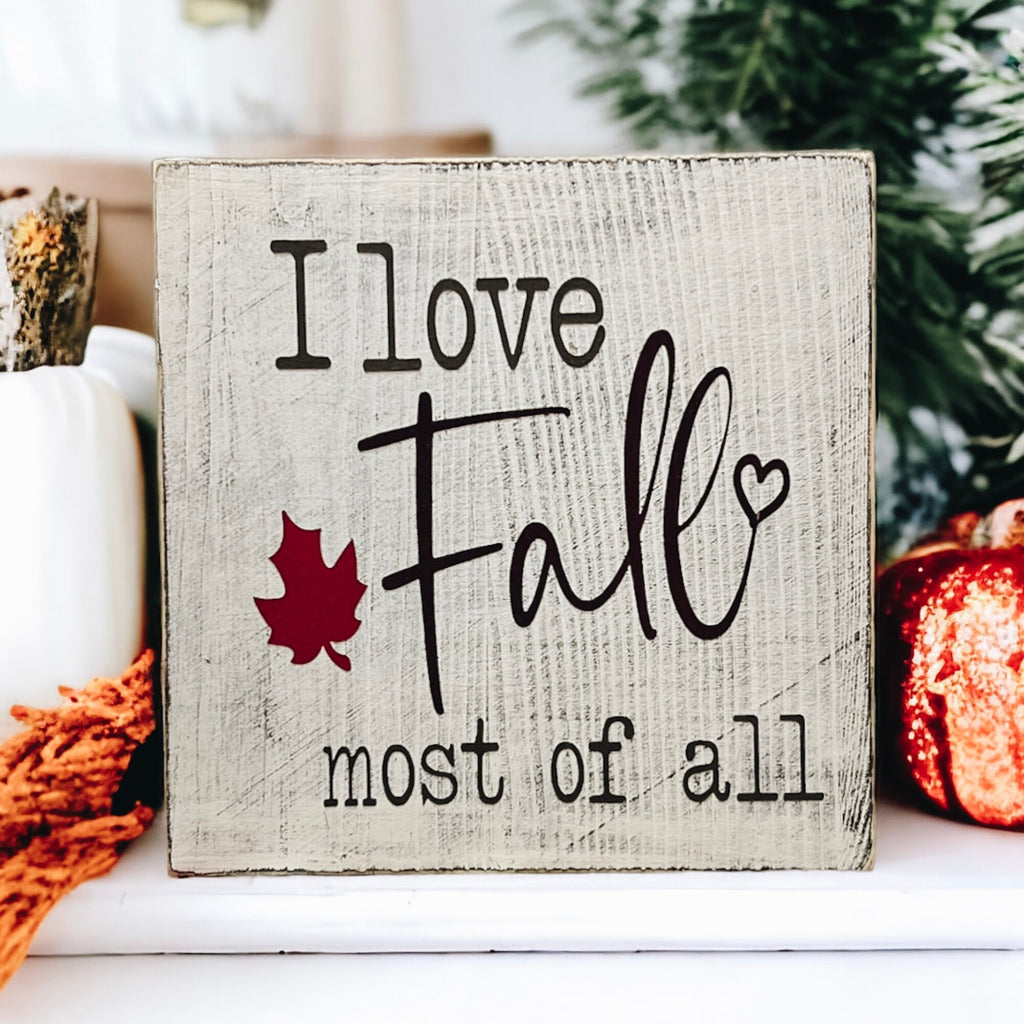 I Love Fall Most of All distressed off-white wood block sign with black lettering and burgundy maple leaf on white mantel with pine cones orange pumpkins and lit pillar candle