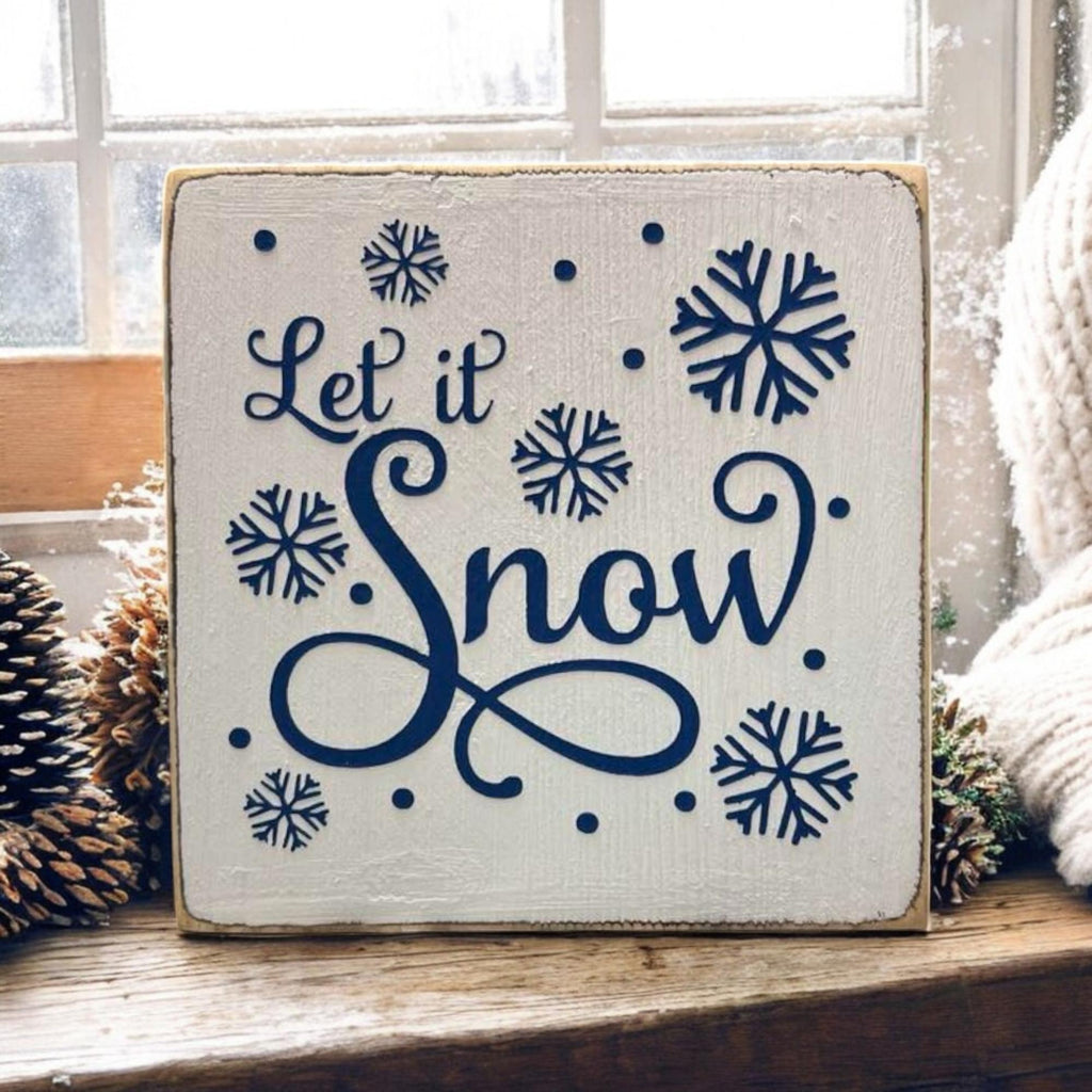 Let It Snow navy wood block sign on snowy windowsill with pine cones and knit scarf