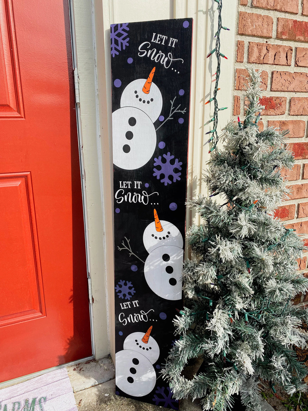 Let It Snow porch leaner sign with blue snowflakes full view leaning against red door with flocked Christmas tree