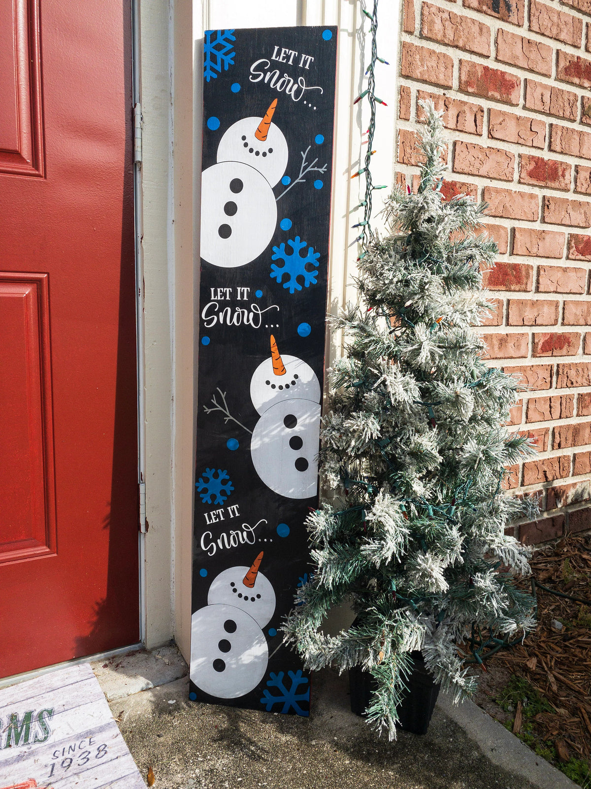 Let It Snow porch leaner sign with purple snowflakes leaning against red front door with flocked Christmas tree