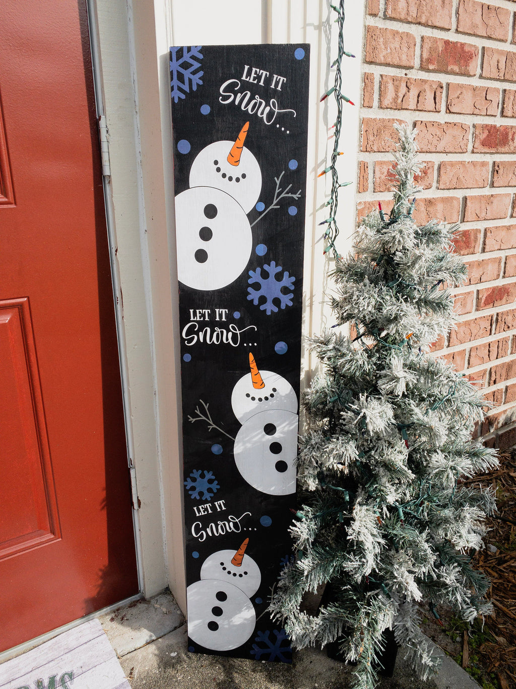 Let It Snow porch leaner sign with blue snowflakes leaning against red front door with flocked Christmas tree