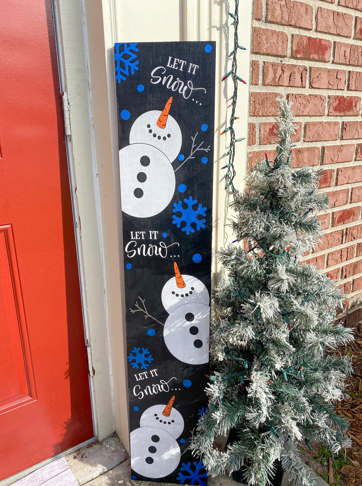 Let It Snow porch leaner sign with blue snowflakes angled view leaning against red front door