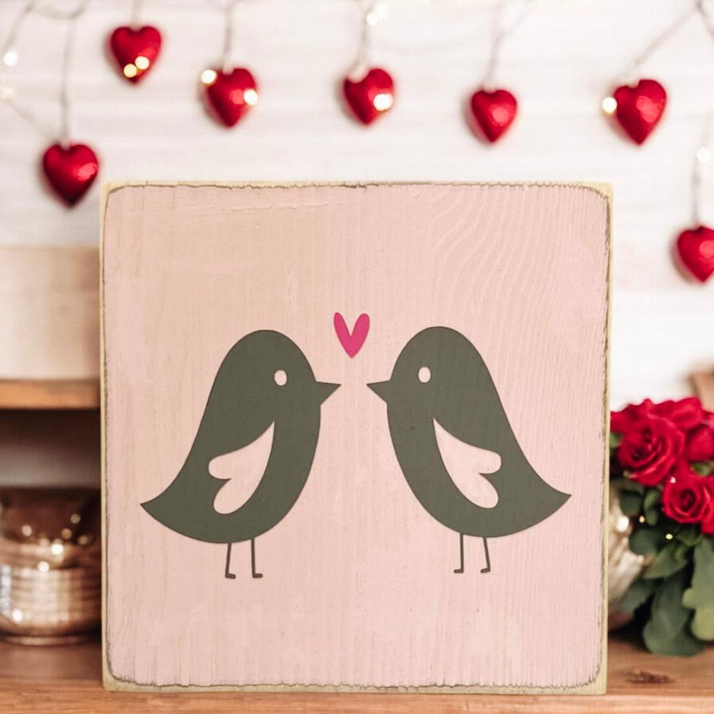 Lovebirds Valentine wood block sign on wood surface with pink rose petals and red heart lights in background