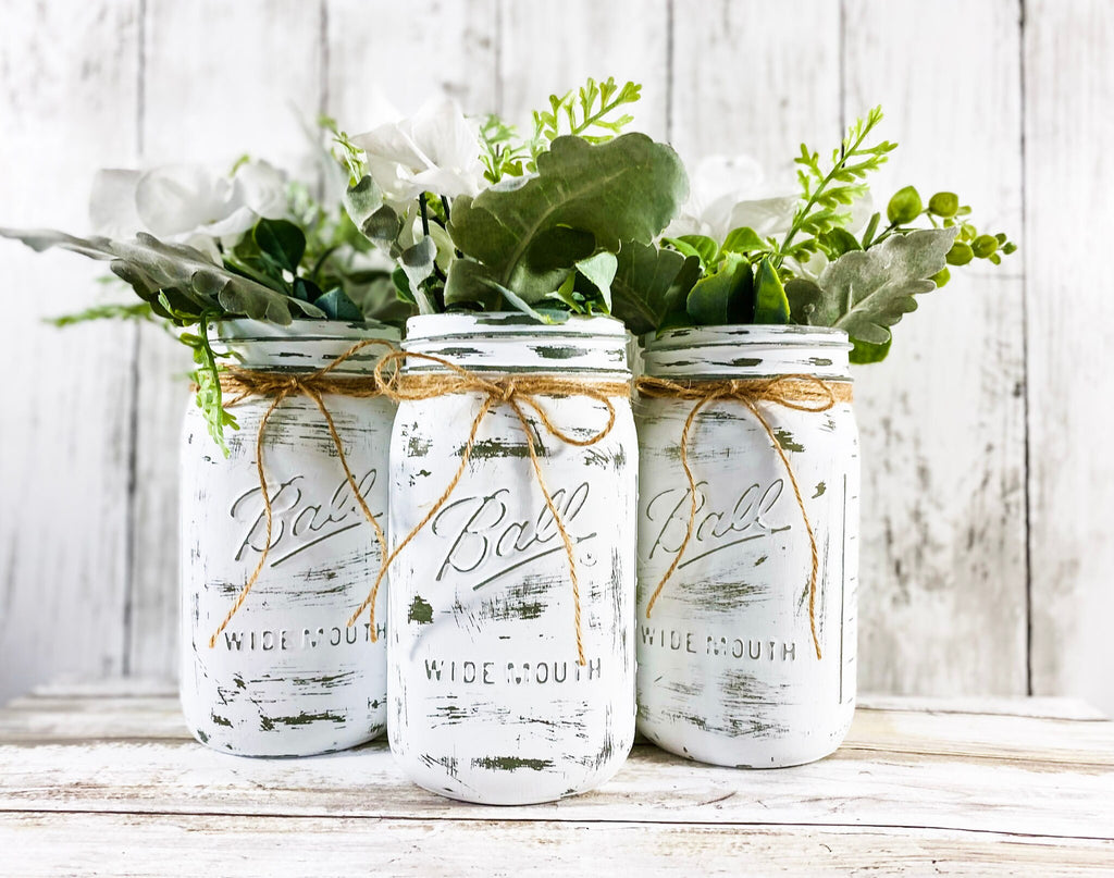 Set of 3 white farmhouse mason jars with greenery styled on white wood surface