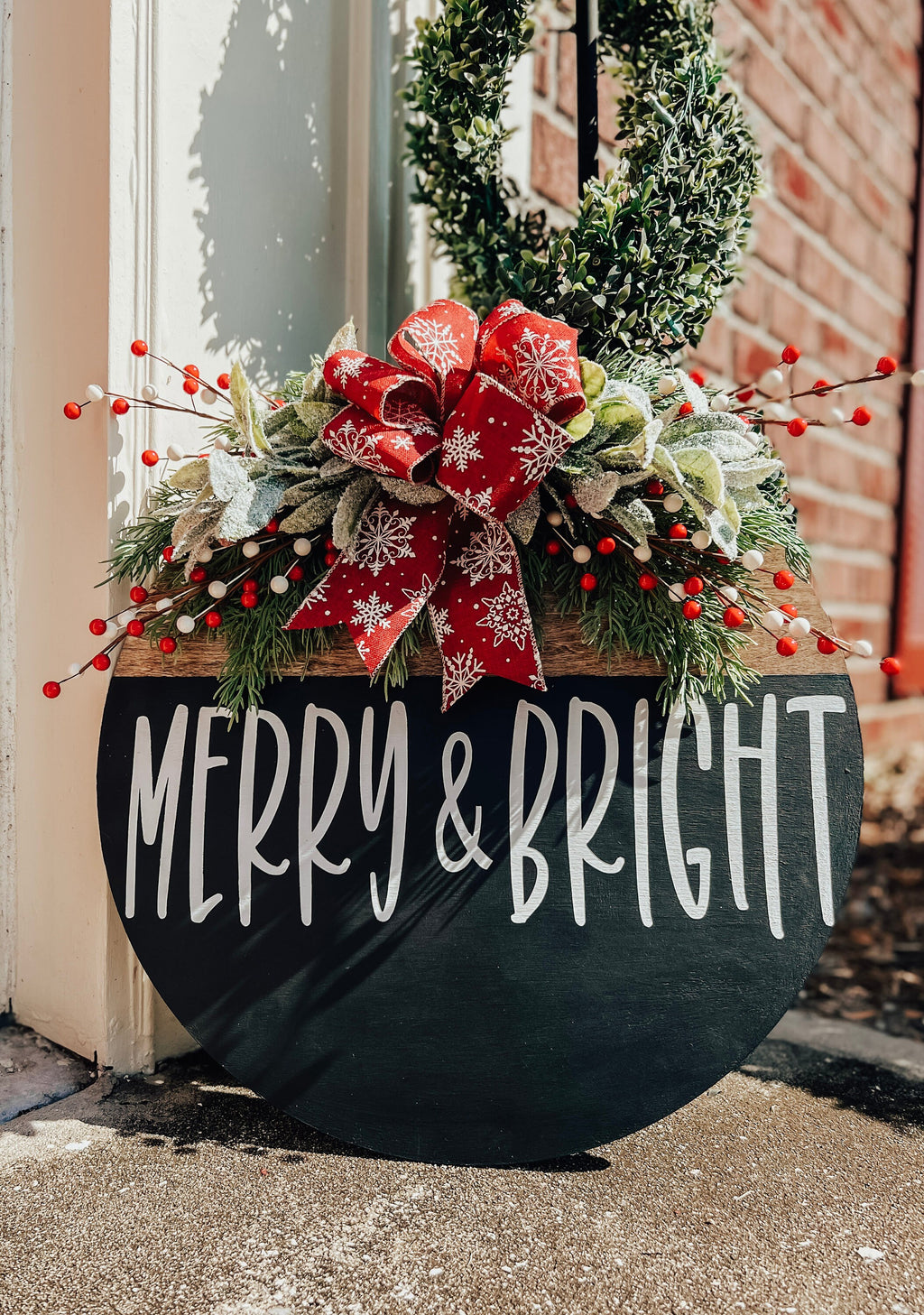Merry and Bright black round wood door hanger with red snowflake bow and frosted greenery leaning against red door on snowy brick porch