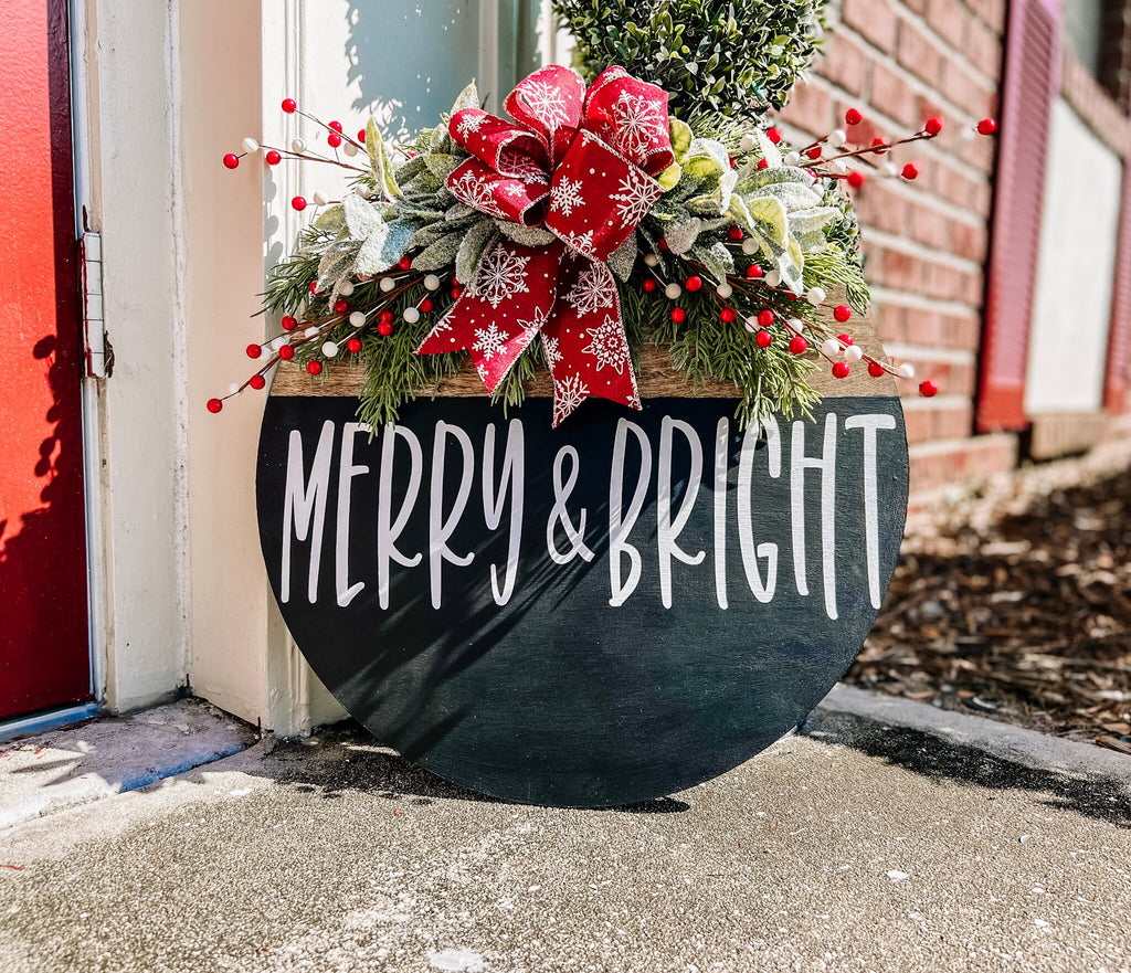 Merry and Bright black round wood door hanger with red snowflake bow and frosted greenery leaning against brick wall with boxwood topiary