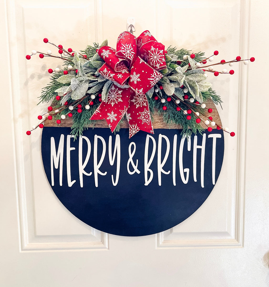 Merry and Bright black round wood door hanger with red snowflake bow and frosted greenery leaning against white door frame on brick porch wide shot