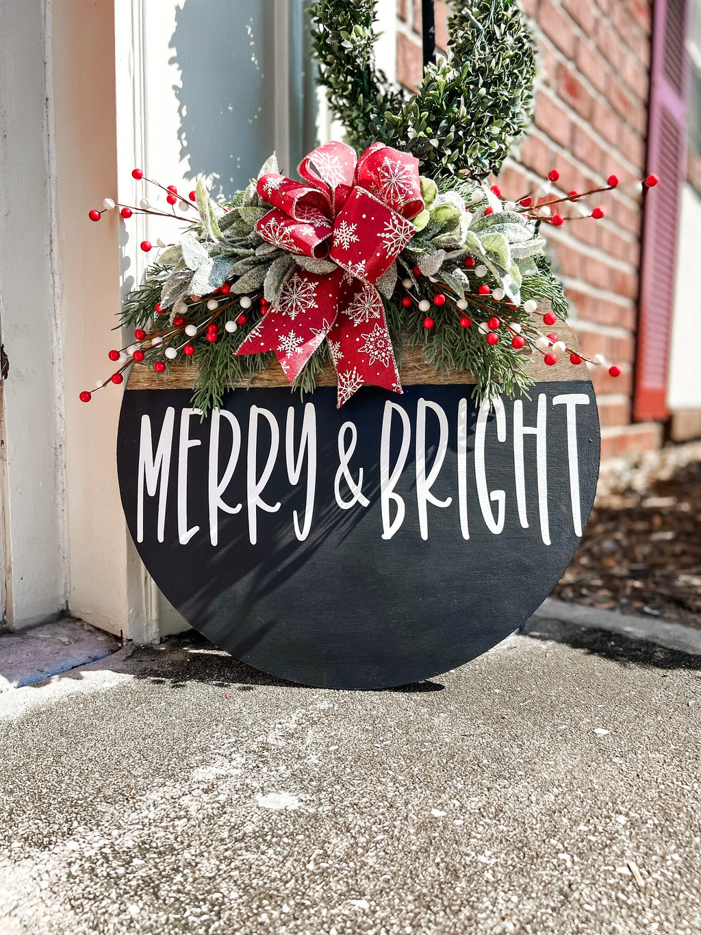 Merry and Bright black round wood door hanger with red snowflake bow and frosted greenery leaning against red door on brick porch angled view