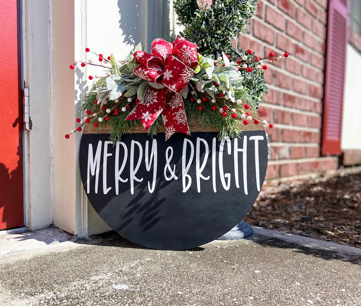 Merry and Bright black round wood door hanger with red snowflake bow and frosted greenery hanging on white paneled door indoors