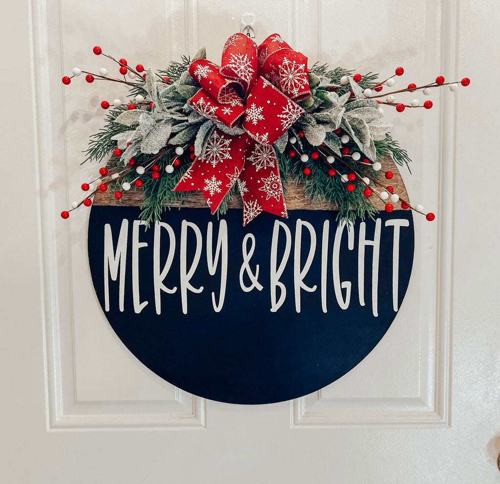 Merry and Bright black round wood door hanger with red snowflake bow and frosted greenery leaning against white door frame on brick porch