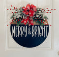 Merry and Bright black round wood door hanger with red snowflake bow and frosted greenery leaning against white door frame on brick porch