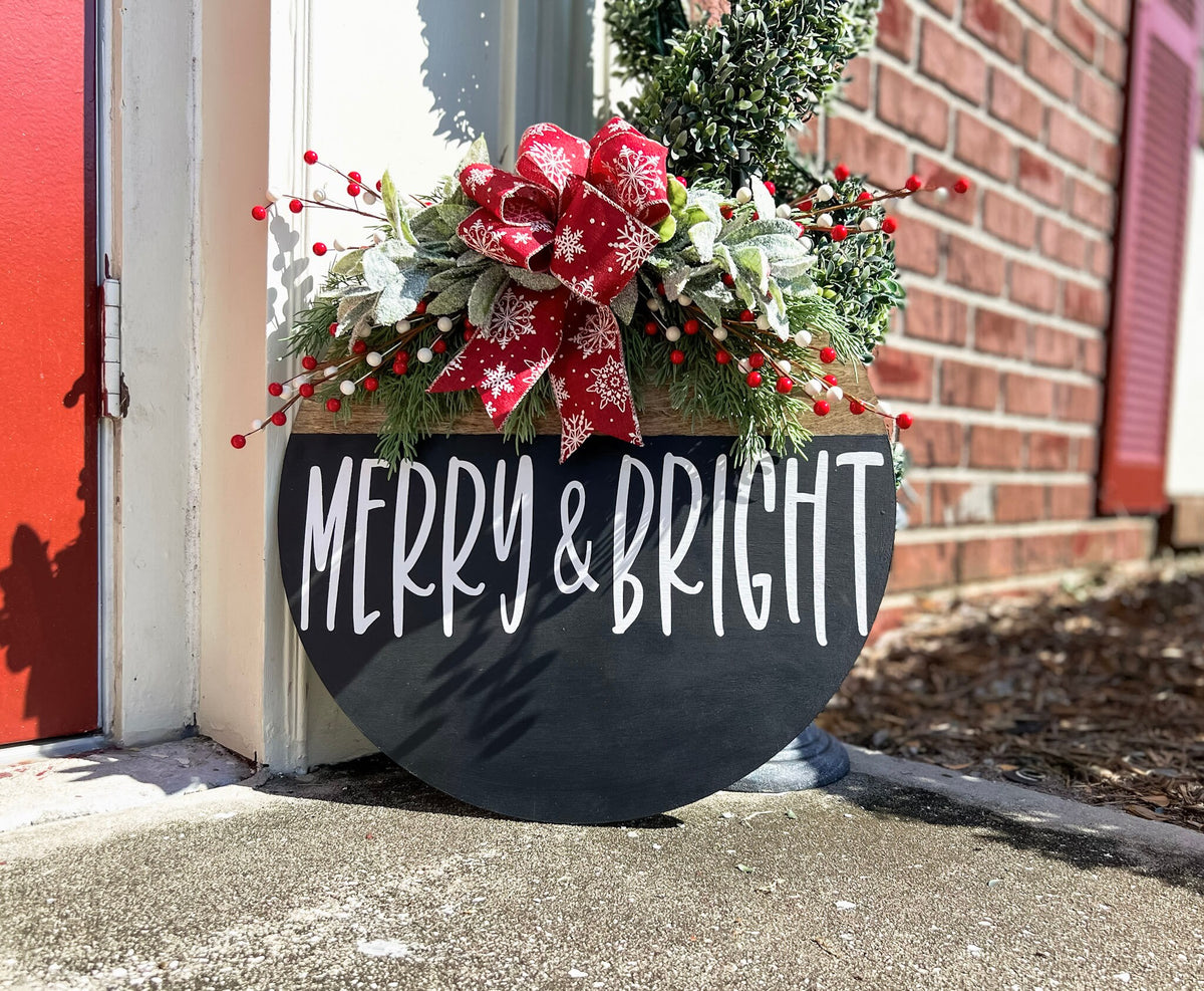Merry and Bright black round wood door hanger with red snowflake bow and frosted greenery leaning against red door on brick porch close up