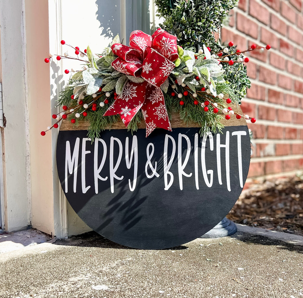 Merry and Bright black round wood door hanger with red snowflake bow and frosted greenery leaning against brick wall with boxwood topiary wide shot