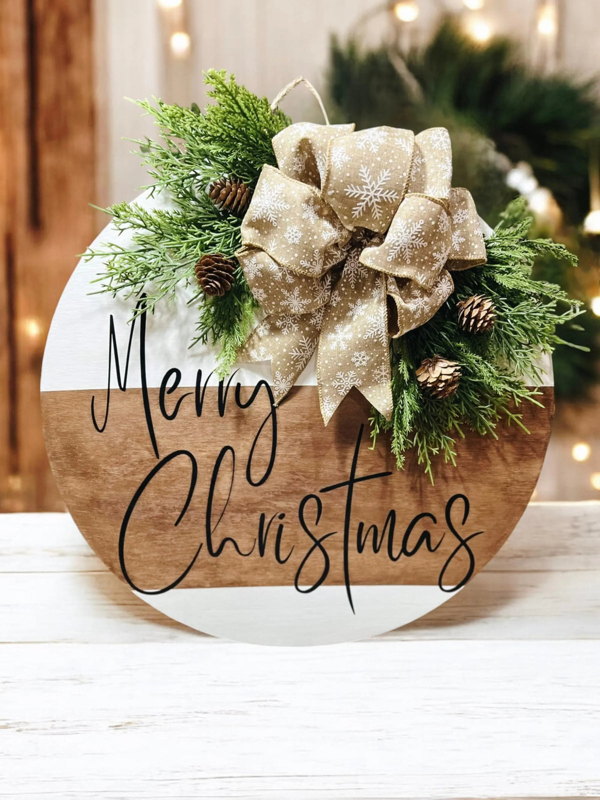 Natural wood and white Merry Christmas round pine door hanger with evergreen branches pinecones and beige snowflake bow hung on rustic white door with warm bokeh string lights