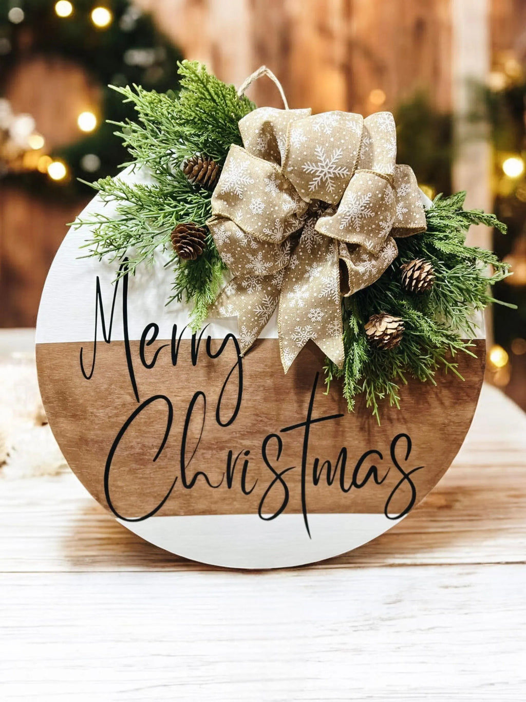 Natural wood and white Merry Christmas round pine door hanger with evergreen branches pinecones and beige snowflake bow on white wood surface with Christmas tree in background angled view
