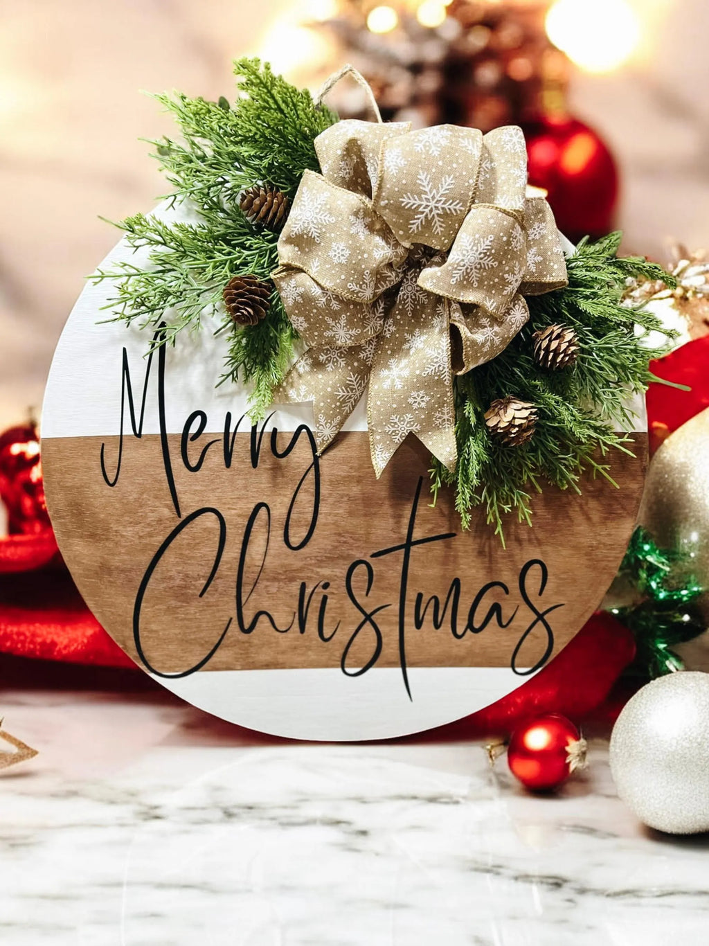 Natural wood and white Merry Christmas round pine door hanger with evergreen branches pinecones and beige snowflake bow on white wood surface with Christmas tree in background wide shot