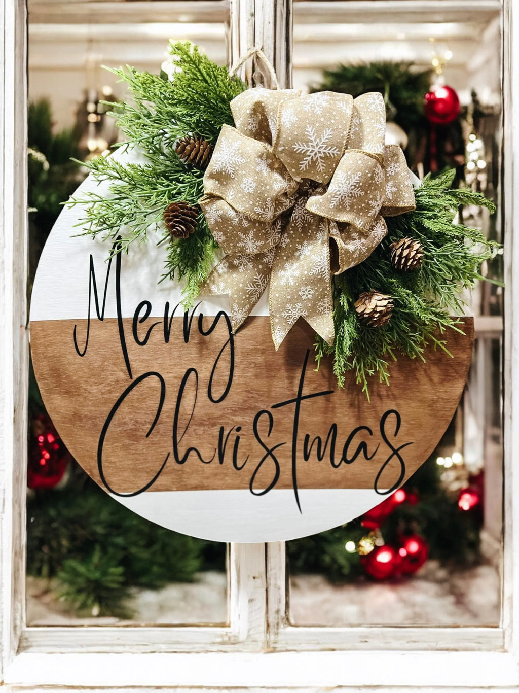 Natural wood and white Merry Christmas round pine door hanger with evergreen branches pinecones and beige snowflake bow on white wood surface with warm bokeh Christmas lights