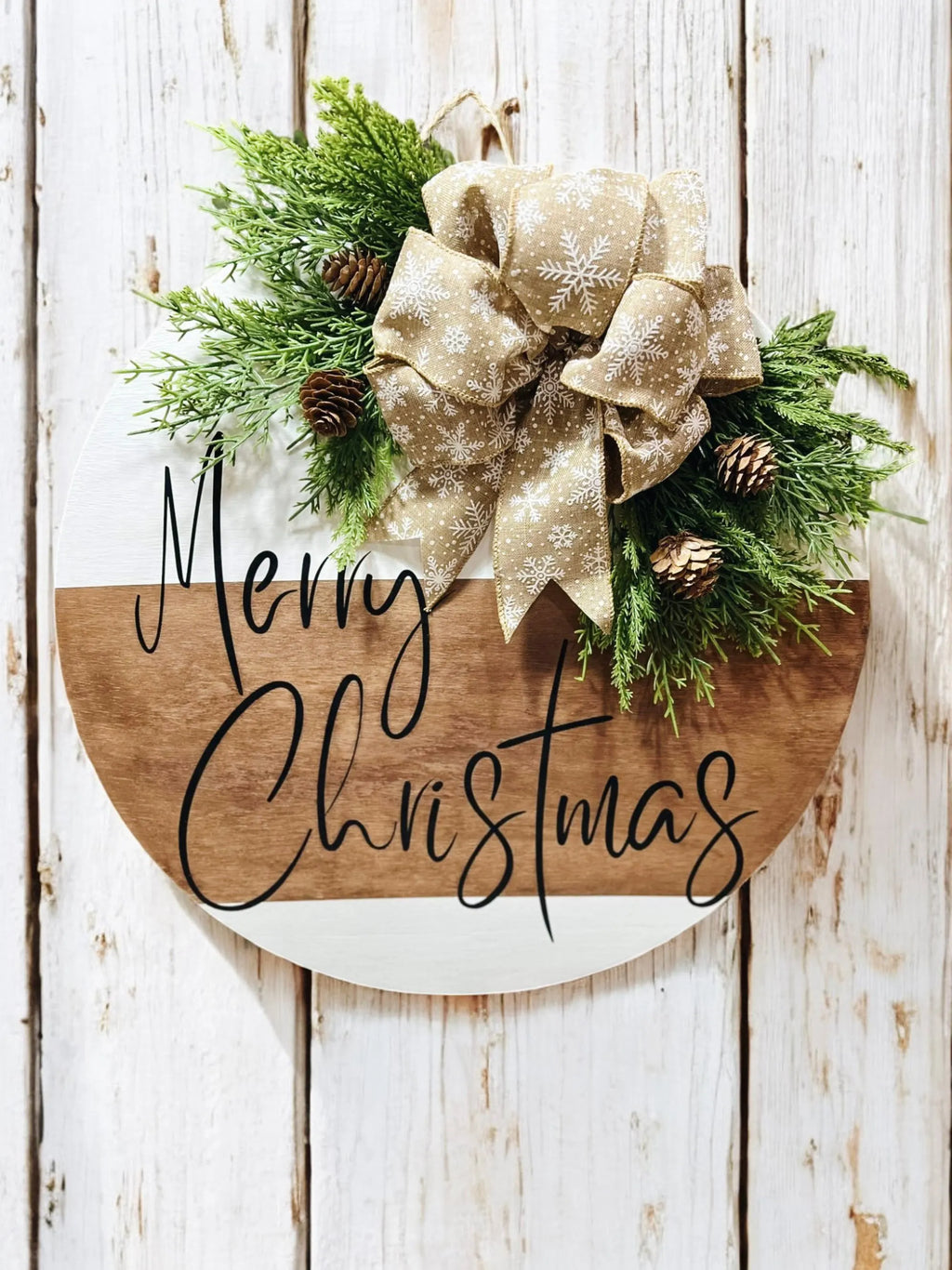 Natural wood and white Merry Christmas round pine door hanger with evergreen branches pinecones and beige snowflake bow on marble surface with red and silver ornaments
