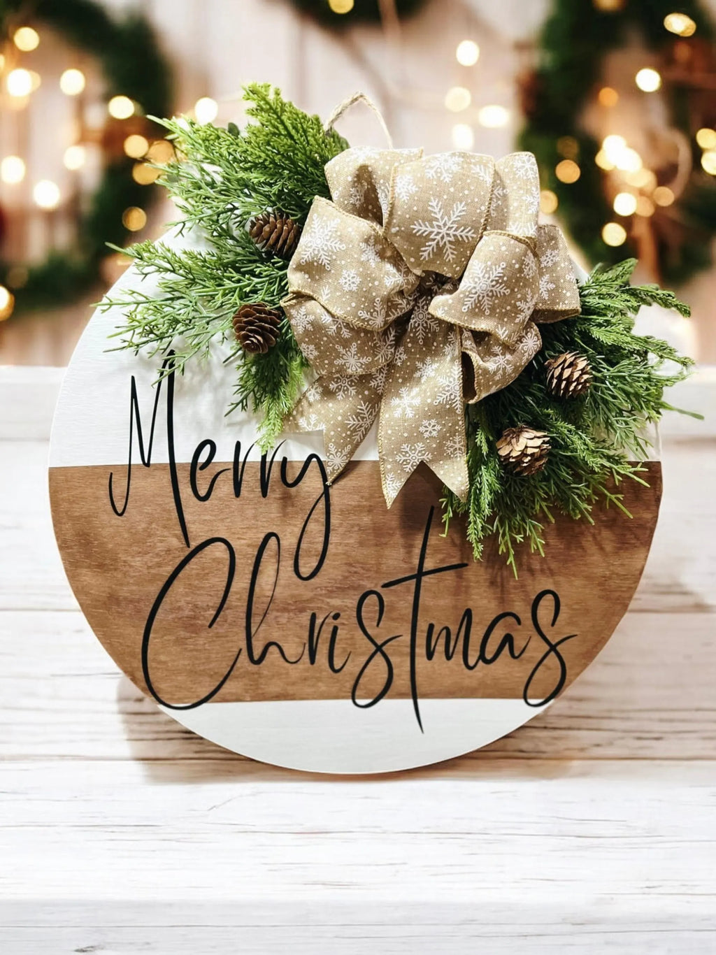 Natural wood and white Merry Christmas round pine door hanger with evergreen branches pinecones and beige snowflake bow hung on rustic white window frame with Christmas tree bokeh