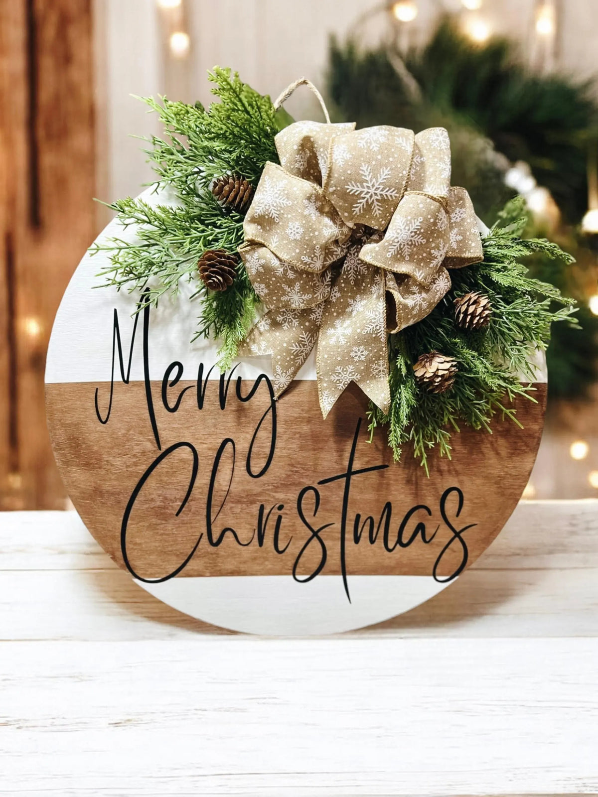 Natural wood and white Merry Christmas round pine door hanger with evergreen branches pinecones and beige snowflake bow hung on white shiplap wall hero shot