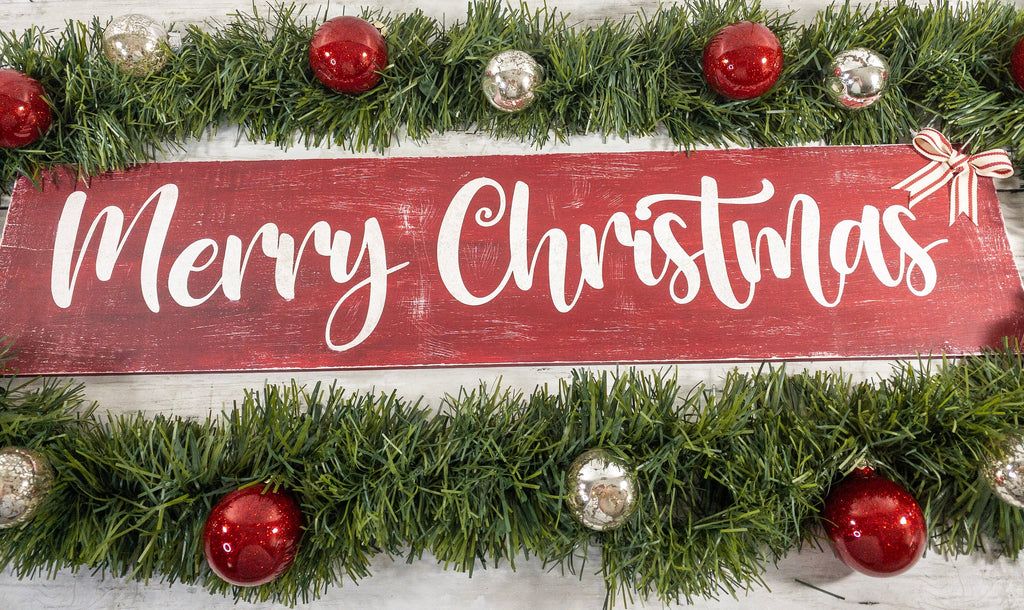 Merry Christmas farmhouse wood sign distressed cranberry red with white script lettering and red white striped bow framed by pine garland with red and silver ornaments on white shiplap close up lettering detail