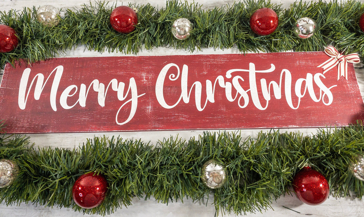 Merry Christmas farmhouse wood sign distressed cranberry red with white script lettering and red white striped bow framed by pine garland with red and silver ornaments on white shiplap close up lettering detail
