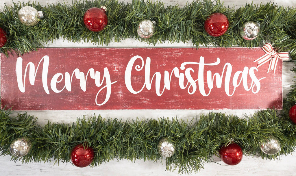 Merry Christmas farmhouse wood sign distressed cranberry red with white script lettering and red white striped bow framed by pine garland with red and silver ornaments on white shiplap angled view
