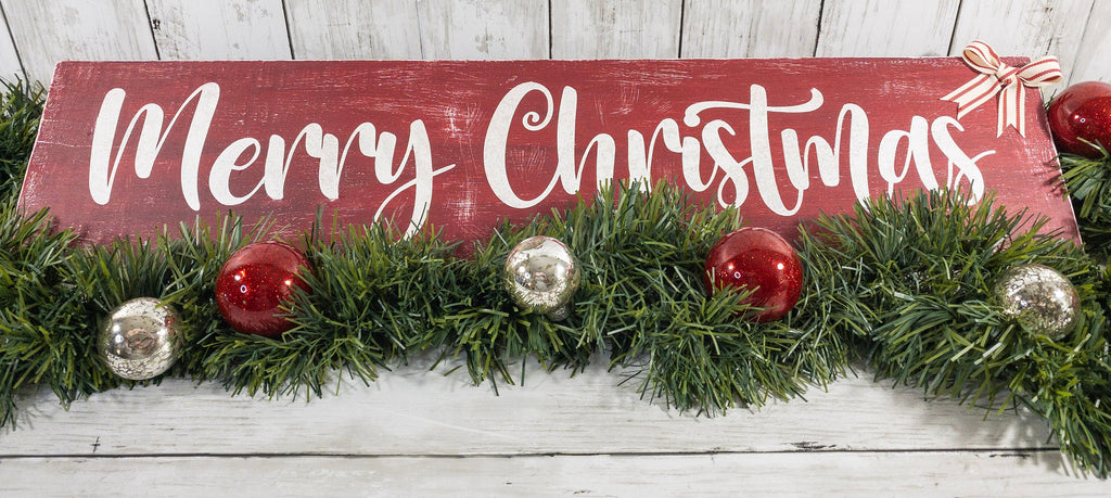Merry Christmas farmhouse wood sign distressed cranberry red with white script lettering and red white striped bow framed by pine garland with red and silver ornaments on white shiplap overhead view