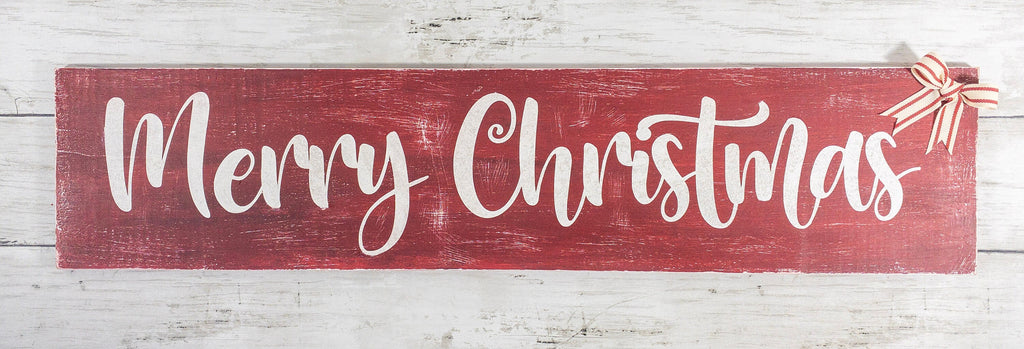 Merry Christmas farmhouse wood sign distressed cranberry red with white script lettering and red white striped bow resting on pine garland with red and silver ornaments below on white shiplap