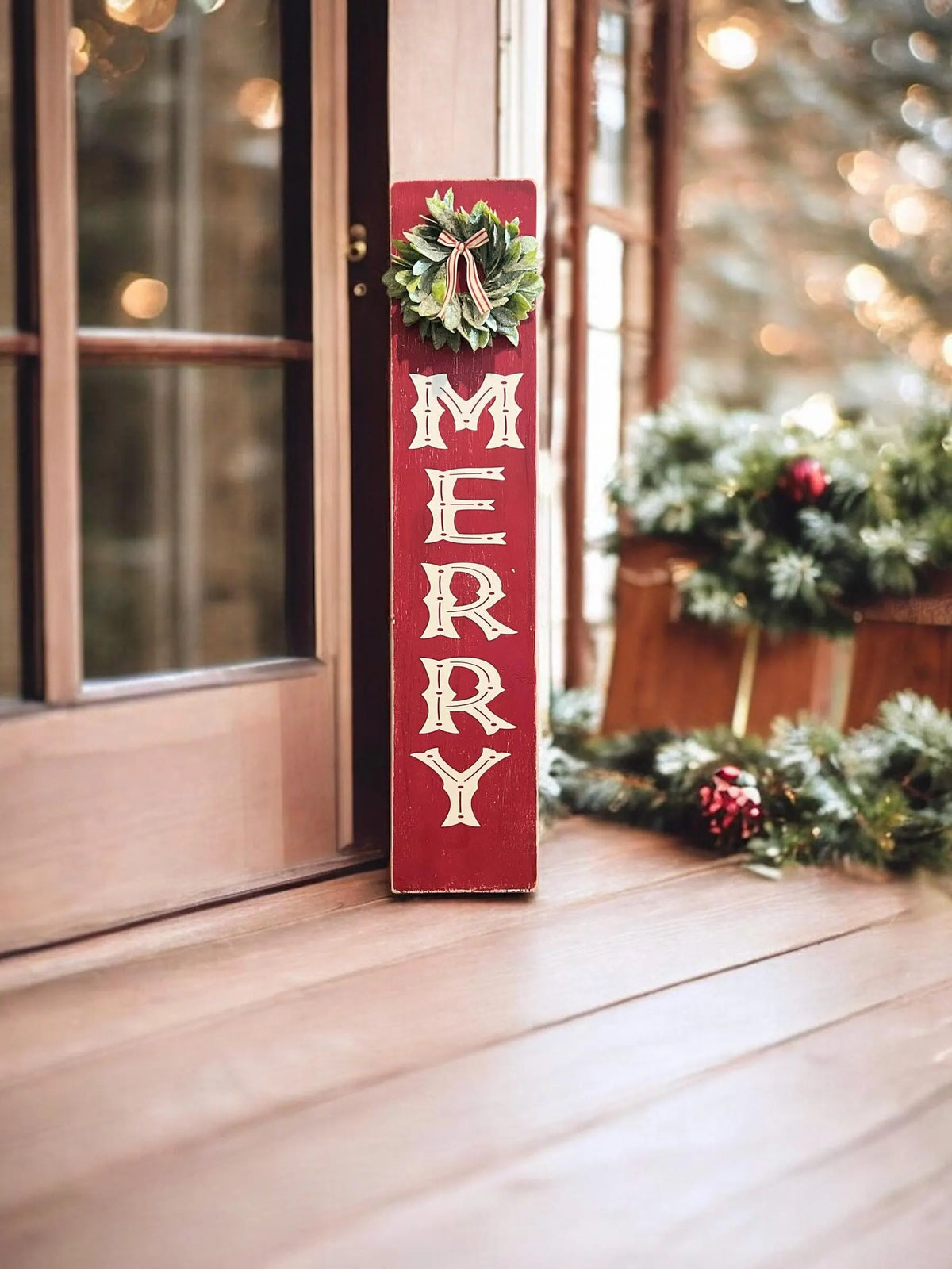 MERRY porch leaner sign with holly wreath leaning against red front door with Christmas trees in pots and lanterns in snow