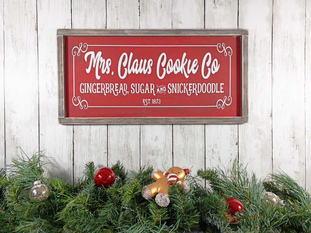 Mrs Claus Cookie Co framed Christmas sign crimson red with white script and serif lettering in distressed gray wood frame on white shiplap with pine garland gingerbread man ornament and red ornaments overhead view