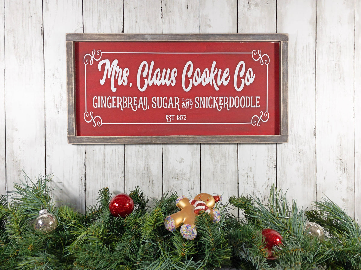 Mrs Claus Cookie Co framed Christmas sign crimson red with white script and serif lettering in distressed gray wood frame on white shiplap with pine garland gingerbread man ornament and red ornaments overhead view
