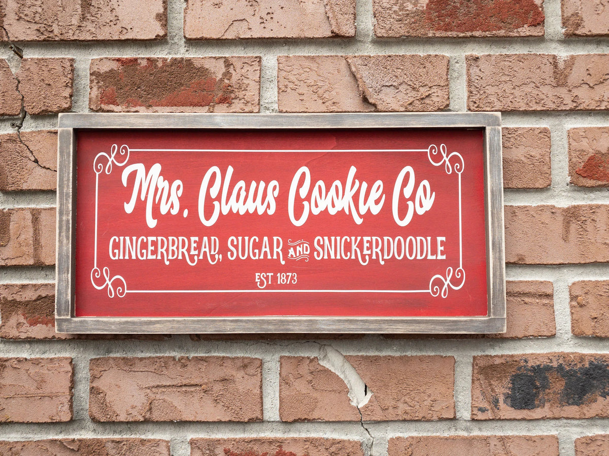 Mrs Claus Cookie Co framed Christmas sign crimson red with white script and serif lettering in distressed gray wood frame on white shiplap with pine garland gingerbread man ornament and red ornaments close up lettering
