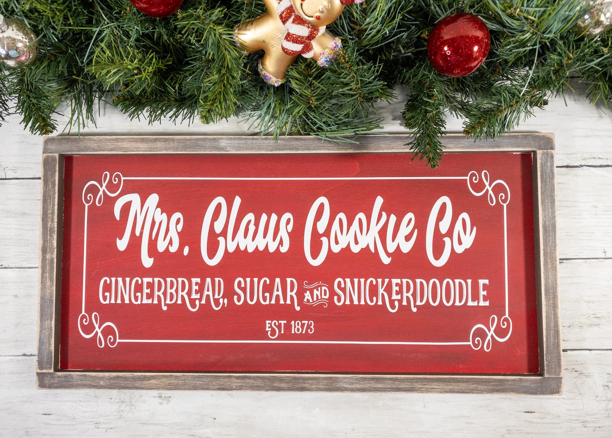 Mrs Claus Cookie Co framed Christmas sign crimson red with white script and serif lettering in distressed gray wood frame on white shiplap with pine garland gingerbread man ornament and red ornaments angled view