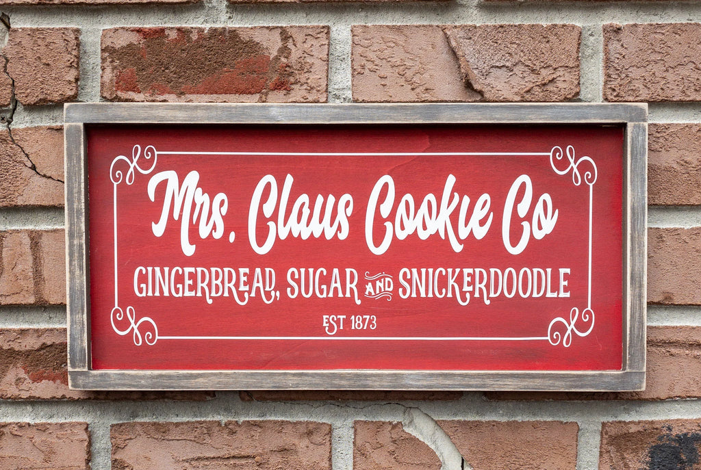 Mrs Claus Cookie Co framed Christmas sign crimson red with white script and serif lettering in distressed gray wood frame on white shiplap with pine garland gingerbread man ornament and red ornaments wide shot