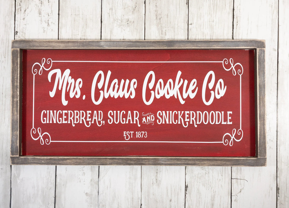 Mrs Claus Cookie Co framed Christmas sign crimson red with white script and serif lettering in distressed gray wood frame mounted on red brick wall wide shot