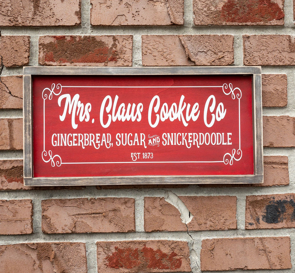 Mrs Claus Cookie Co framed Christmas sign crimson red with white script and serif lettering in distressed gray wood frame on white shiplap with pine garland gingerbread man ornament and red ornaments below