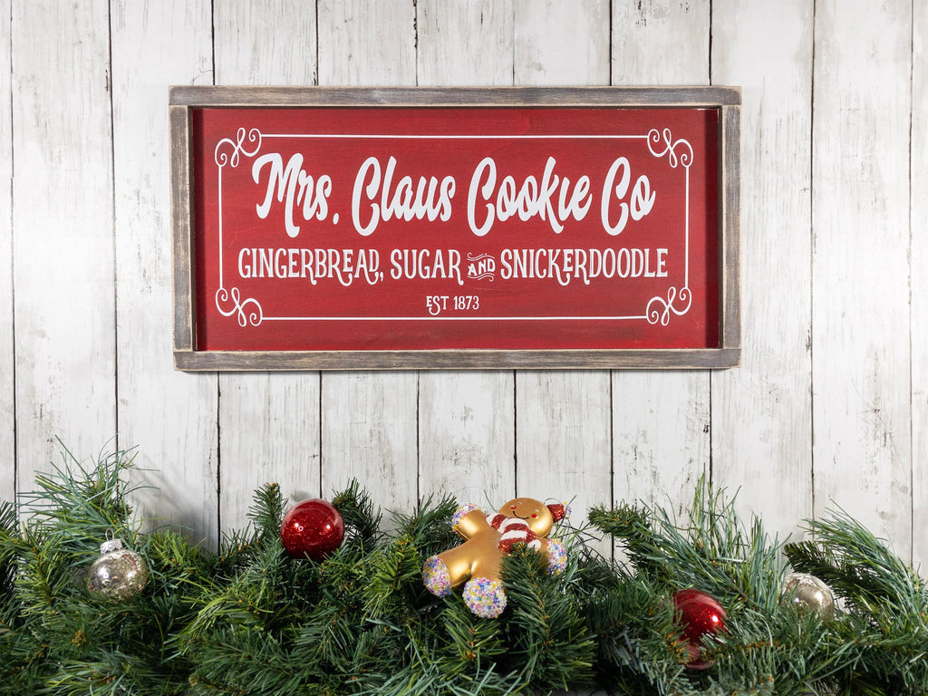 Mrs Claus Cookie Co framed Christmas sign crimson red with white script and serif lettering in distressed gray wood frame mounted on red brick wall