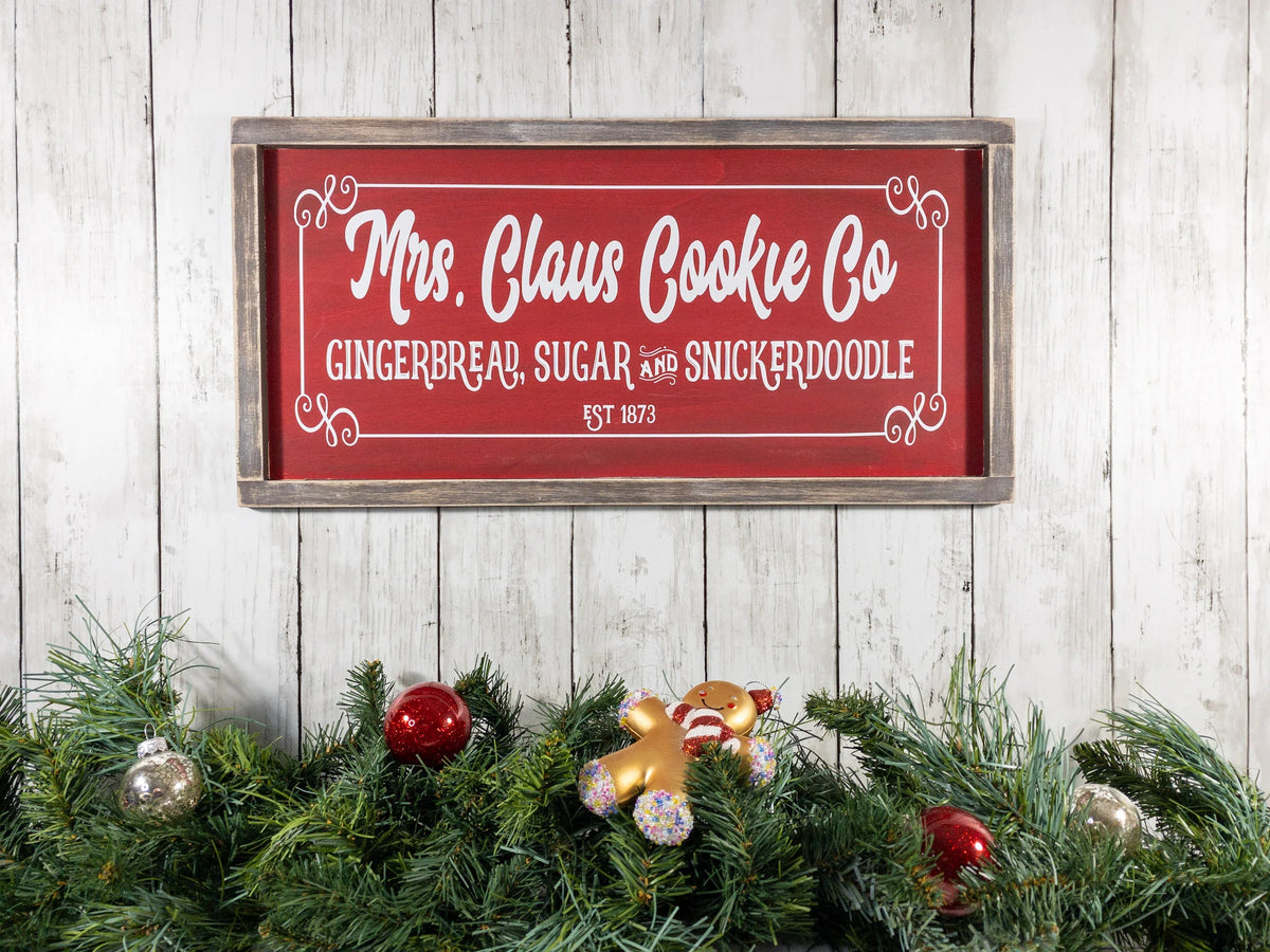 Mrs Claus Cookie Co framed Christmas sign crimson red with white script and serif lettering in distressed gray wood frame mounted on red brick wall
