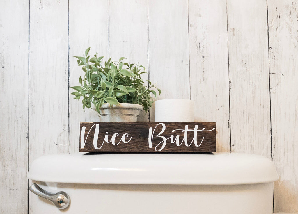 Nice Butt dark stained farmhouse toilet paper box on white surface with plant and toilet paper roll styled view